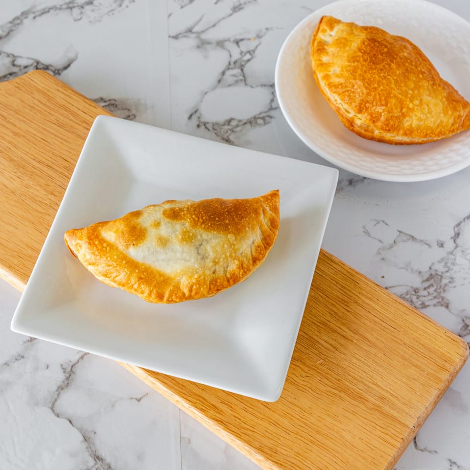 Two golden-brown Argentinian empanadas, one on a white square plate, another on a round plate.