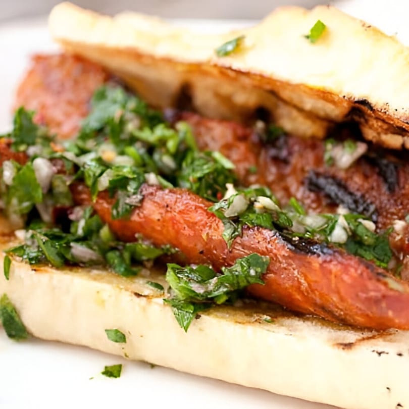 Close-up of a choripán, an Argentinian chorizo sandwich, topped with vibrant green chimichurri sauce.