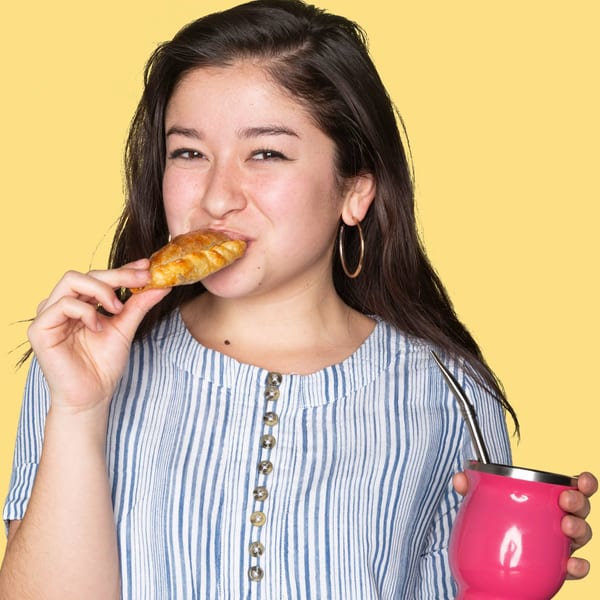 Smiling woman in a striped shirt eating an Argentinian empanada and holding a pink mate cup.