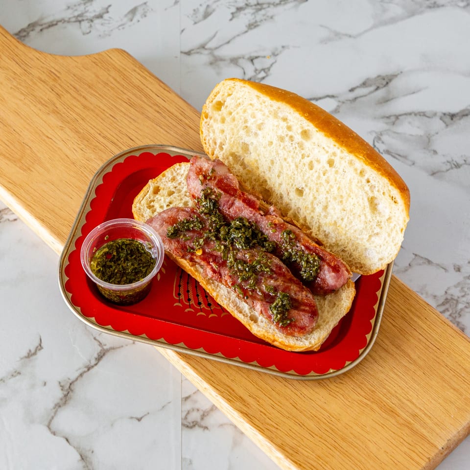 Two chorizos with chimichurri on a baguette, served on a red tray with a side of chimichurri.