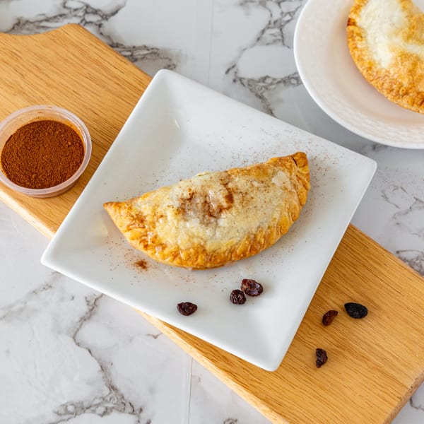 A golden-brown Argentinian empanada dusted with cinnamon and sugar, served on a white plate with raisins.