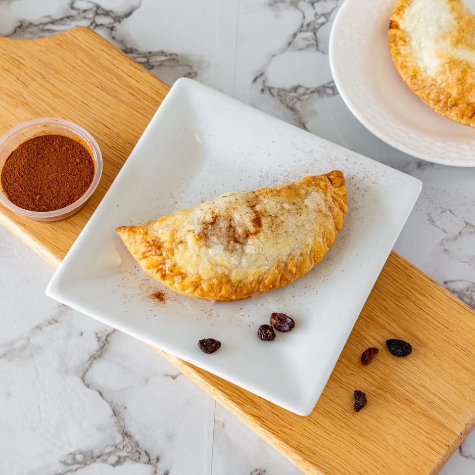 A golden-brown Argentinian empanada dusted with cinnamon and sugar, served on a white plate with raisins.