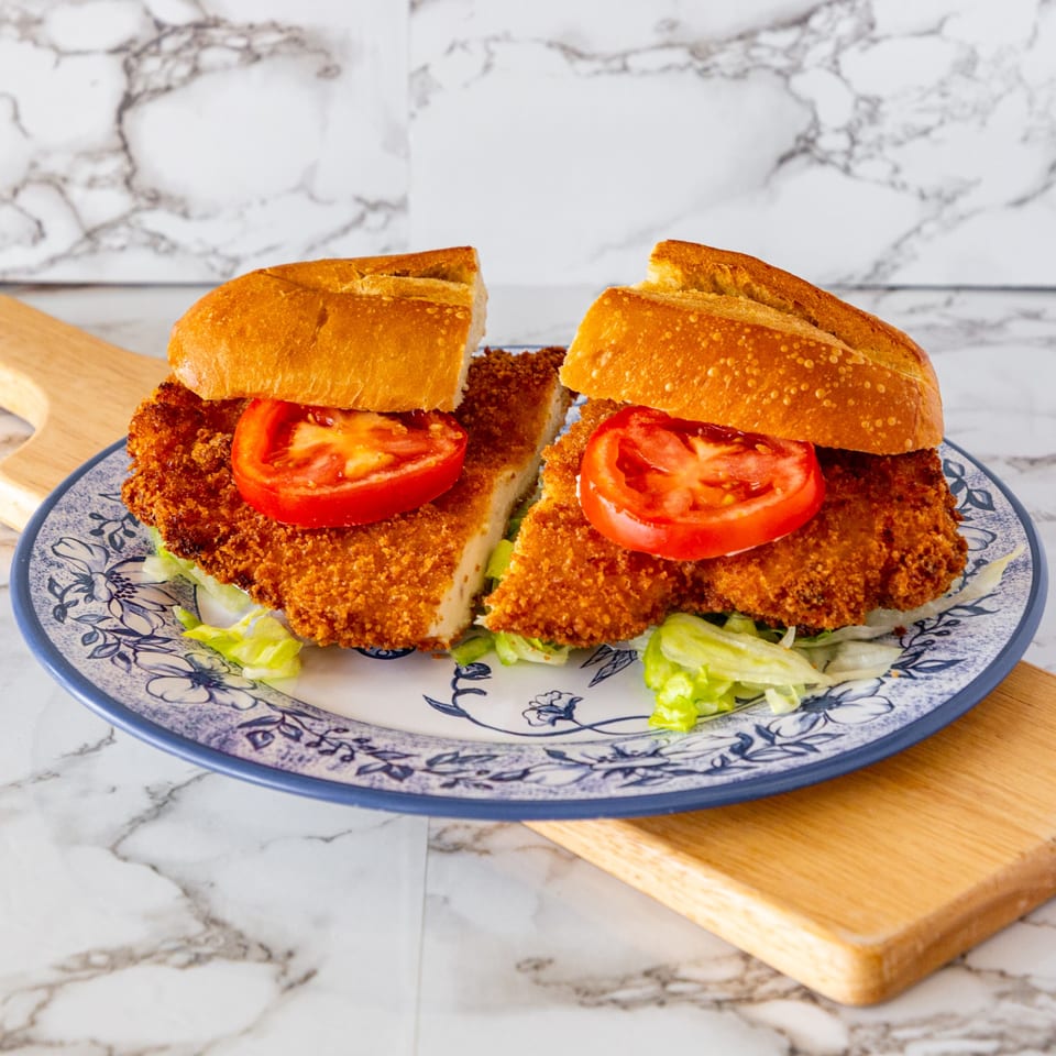 Two halves of a milanesa sandwich with lettuce and tomato on a blue and white plate.