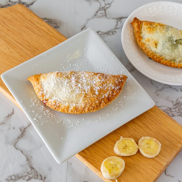 A golden-brown Argentinian empanada dusted with powdered sugar on a white plate, with banana slices.