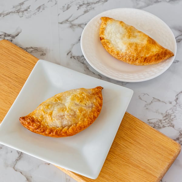 Two golden-brown Argentinian empanadas, one on a white rectangular plate, the other on a round plate.