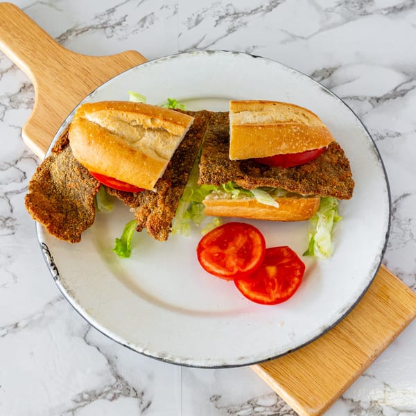 Two halves of a milanesa sandwich with lettuce and tomato, on a white plate.